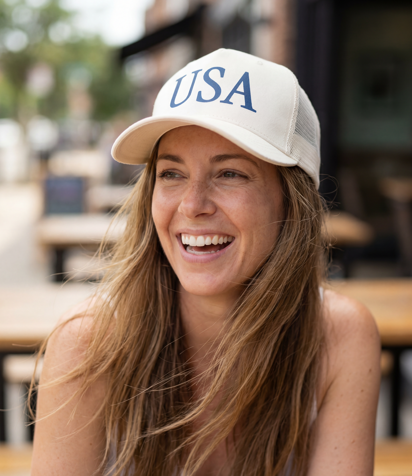 The Alex Jones Store - Smiling brunette woman wearing white 'USA' Premium Trucker Hat with navy serif USA print, outdoor cafe setting